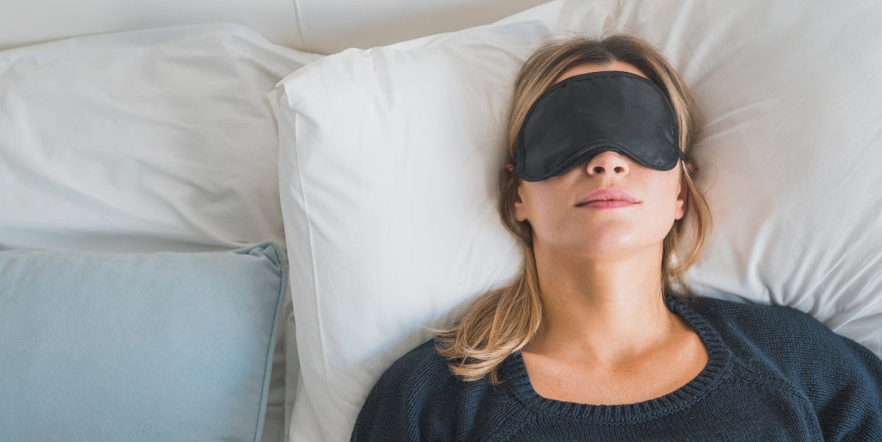 A woman wearing a black sleep mask lies in bed with white pillows and bedding, covered with a white blanket, next to a light blue pillow.
